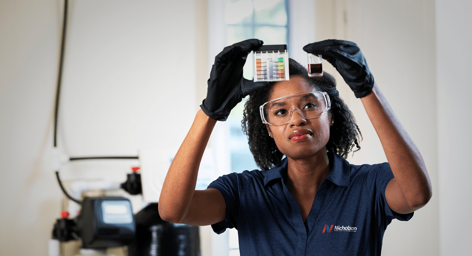 A Nicholson technician in a blue uniform and safety gear compares a water sample to a testing chart in front of a water softener system.