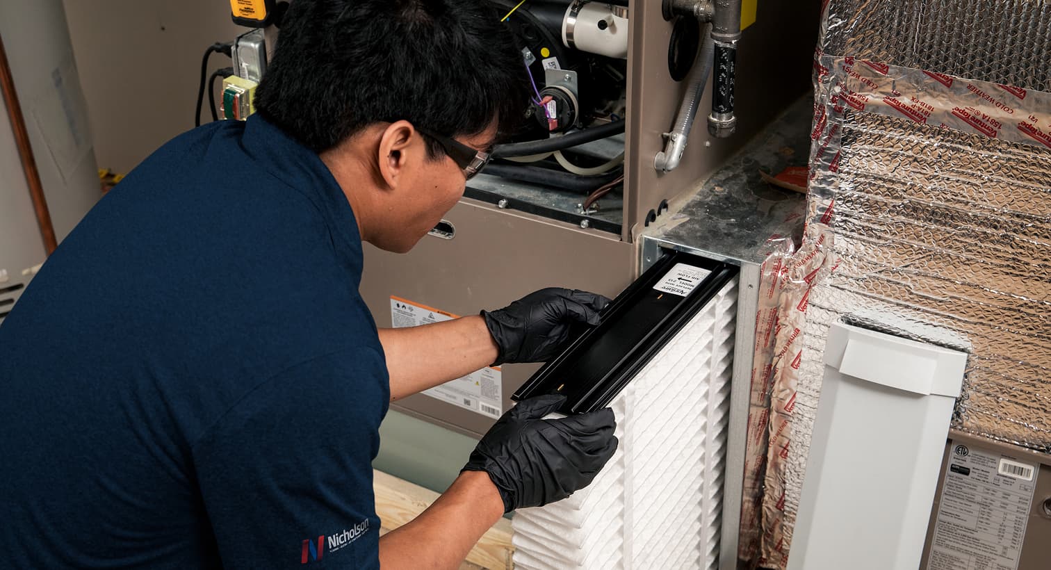 A professional Nicholson HVAC technician replacing a clean pleated air filter in an indoor air handler unit during a routine AC maintenance tune-up.