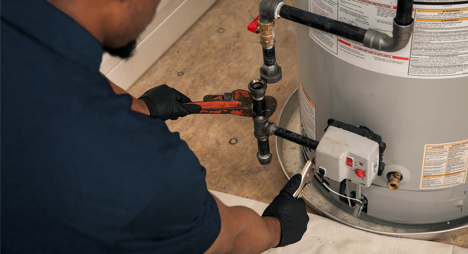A Nicholson plumber wearing black gloves uses a pipe wrench to repair the gas line and control valve assembly on a residential water heater tank.